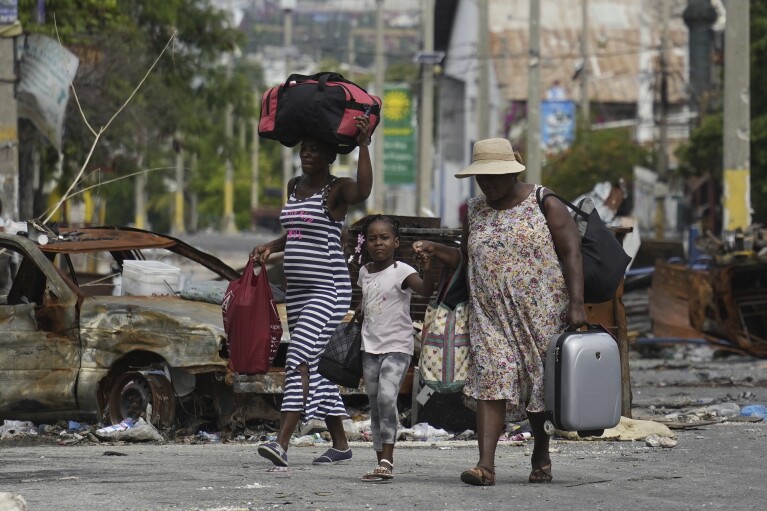 FILE - A little girl is led by the hand past a burnt car blocking the street as residents evacuate the Delmas 22 neighborhood to escape gang violence in Port-au-Prince, Haiti, Thursday, May 2, 2024. As young Haitians are increasingly exposed to violence, the country is undergoing a wider push to dispel a long-standing taboo on seeking therapy and talking about mental health. (AP Photo/Ramon Espinosa, File)