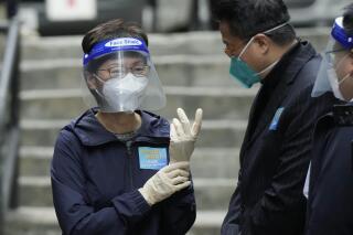 La jefa territorial de Hong Kong, Carrie Lam (izquierda), se coloca unos guantes antes de entregar paquetes con materiales para la prevención del coronavirus a residentes durante un acto en Hong Kong, el 2 de abril de 2022. (AP Foto/Kin Cheung)