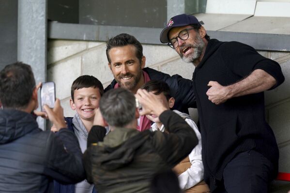 Wrexham Co-Owner, Ryan Reynolds, center, and Hugh Jackman, right, pose for a photo with a fans in the stands before the Sky Bet League Two soccer match between Wrexham and Milton Keynes Dons at the SToK Racecourse, Wrexham, Wales, Saturday Aug. 5, 2023. (Jacob King/PA via AP)