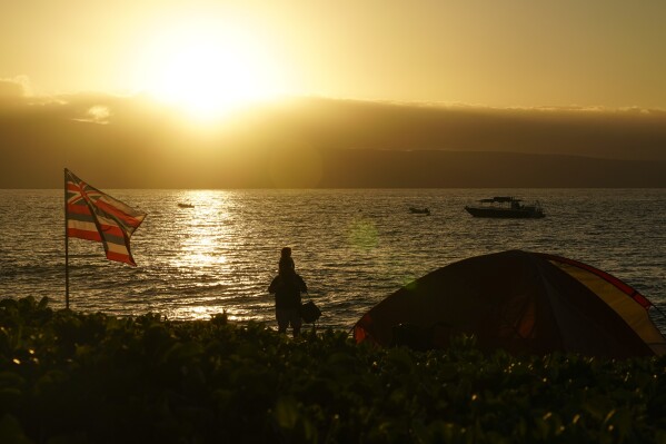 A child sits atop a person's shoulders next to a flag of Hawaii and a tent on Kaanapali Beach, Wednesday, Dec. 6, 2023, in Lahaina, Hawaii. A group of survivors is camping on the resort beach to protest and raise awareness for better long-term housing options for those displaced. Residents and survivors still dealing with the aftermath of the August wildfires in Lahaina have mixed feelings as tourists begin to return to the west side of Maui, staying in hotels still housing some displaced residents. (AP Photo/Lindsey Wasson)