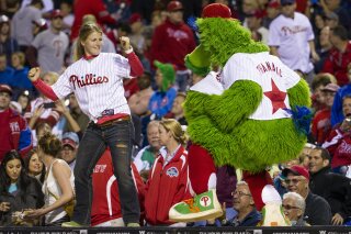 File-This Sept. 16, 2013. file photo shows the Phillies Phanatic dancing with a fan on the dugout during the eighth inning of a baseball game in Philadelphia. The Philadelphia Phillies have sued the New York company that created the Phanatic mascot to prevent the green fuzzy fan favorite from becoming a free agent. In a complaint filed Friday, Aug. 2, 2019, in U.S. District Court in Manhattan, the team alleged Harrison/Erickson threatened to terminate the Phillies' rights to the Phanatic next year and “make the Phanatic a free agent” unless the team renegotiated its 1984 agreement to acquire the mascot’s rights. (AP Photo/Chris Szagola, File)