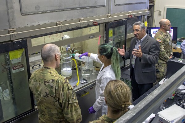 This September 2022, image provided by Los Alamos National Laboratory shows Lt. Gen. Thomas Bussiere, left, and Associate Laboratory Director for Weapons Engineering James Owen, right, watch as scientist Bi Nguyen demonstrates the wet slurry formulation process in Los Alamos, N.M. Owen said the national security work done at the lab is crucial as global instability mounts. (Los Alamos National Laboratory via AP)
