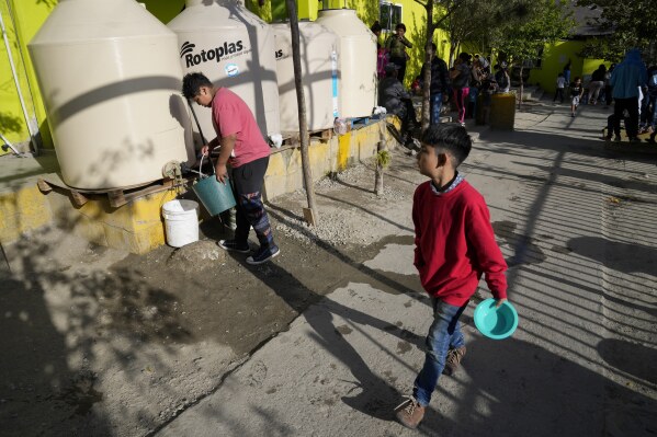 A migrant fills up a bucket with water from a cistern as a boy passes at the Agape migrant shelter Tuesday, May 9, 2023, in Tijuana, Mexico. Among the last cities downstream to receive water from the shrinking Colorado River, Tijuana is staring down a water crisis. (AP Photo/Gregory Bull)