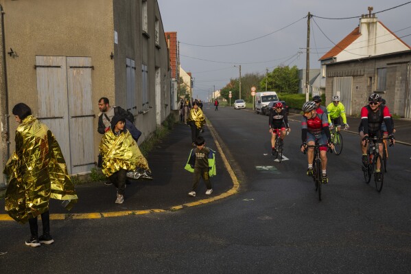 As cyclists ride by, a group of Kurdish migrants from Iran and Iraq who were unsuccessful in their attempt to reach the United Kingdom by boat walk back to the town of Ambleteuse in northern France, on Sunday, May 19, 2024 after being discovered by the police. (AP Photo/Bernat Armangue)