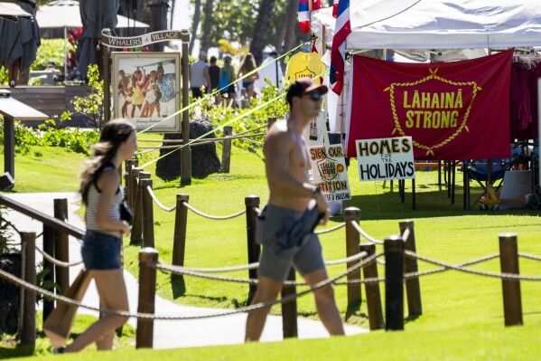 FILE - People walk by a tent with a "Lahaina Strong" flag, Wednesday, Dec. 6, 2023, at Whalers Village on Kaanapali Beach in Lahaina, Hawaii, as a group of wildfire survivors camps on the resort beach to protest and raise awareness for better long-term housing options for those displaced. (AP Photo/Lindsey Wasson, File)