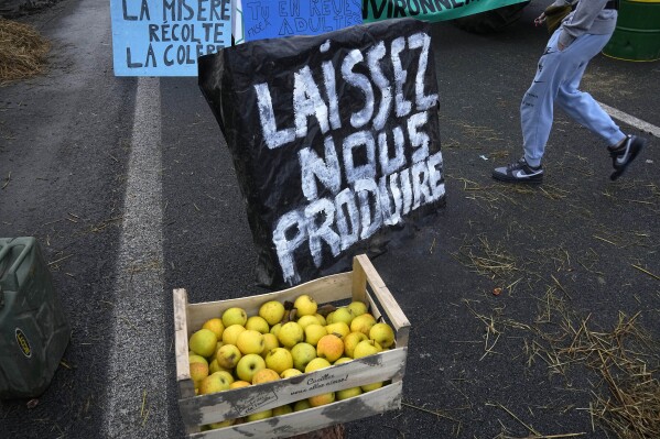 FILE - Apples are made available next to a poster reading " Let us produce" as farmers block highway, on Feb. 1, 2024 in Argenteuil, north of Paris. European farmers and households are both hurting these days because of multiple factors, including persistent inflation, high interest rates and volatile energy prices. (AP Photo/Michel Euler, File)
