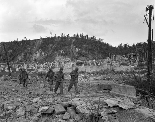 FILE - In this Aug. 9, 1944 file photo, U.S. soldiers walk by a bombed out cemetery in Agana, Guam. The 1941 Japanese invasion of Guam, which happened on the same December day as the attack on Hawaii's Pearl Harbor, set off years of forced labor, internment, torture, rape and beheadings.  Now, more than 75 years later, thousands of people on Guam, a U.S. territory, are expecting to get long-awaited compensation for their suffering at the hands of imperial Japan during World War II. (AP Photo/Joe Rosenthal, File)