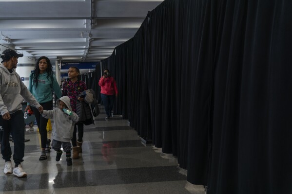 Run by a private firm hired by the city, people walk next to a makeshift shelter hidden by a long row of black curtains at O'Hare International Airport, Wednesday, Sept. 20, 2023, in Chicago. Unlike migrants in the public eye at police stations, the migrants at O'Hare and a handful at Midway International Airport have limited access to resources, including showers and medical care. (AP Photo/Erin Hooley)