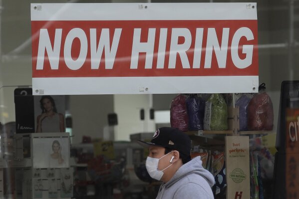 FILE - This May 7, 2020, file photo shows a man wearing a mask while walking under a Now Hiring sign at a CVS Pharmacy during the coronavirus outbreak in San Francisco.  Friday, Dec. 4, monthly U.S. jobs report will help answer a key question hanging over the economy: Just how much damage is being caused by the resurgent coronavirus, the resulting restrictions on businesses and the reluctance of consumers to shop, travel and dine out?  (AP Photo/Jeff Chiu, File)