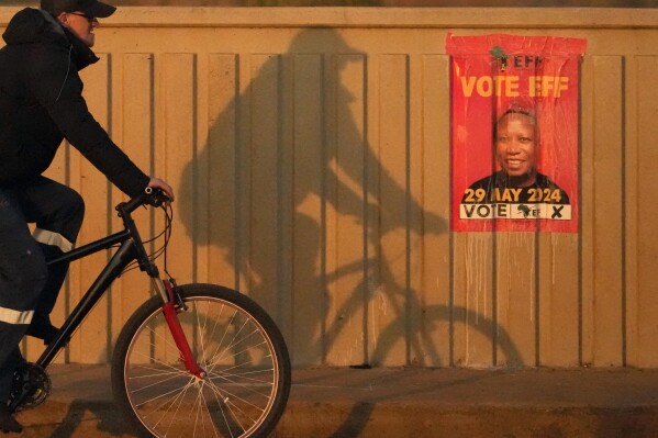 2024年5月27日，星期一，在南非约翰内斯堡以东的贝诺尼，一名男子骑自行车经过一张经济自由战士（EFF）海报，海报上是该组织领导人朱利叶斯·马莱马（Julius Malema）的脸。在2024年5月29日星期三的主要选举之前，获得提前投票特别许可的南非人星期一进行了投票。（美联社照片/Themba Hadebe）