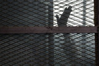 FILE - In this Aug. 22, 2015 file photo, a Muslim Brotherhood member waves his hand from a defendants cage in a courtroom in Torah prison, southern Cairo, Egypt. A leading human rights group says the coronavirus has struck several Egyptian prisons and killed several detainees, as authorities seek to stifle news of the virus’s spread behind bars. Human Rights Watch, released an extensive report Monday, July 20, 2020 documenting multiple cases of detainees who died after experiencing virus symptoms without being tested or receiving adequate medical treatment. (AP Photo/Amr Nabil, File)