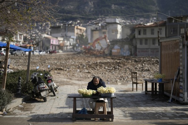 A man displays flowers for sale next to remains of destroyed buildings in the city center of Antakya, southern Turkey, Thursday, Jan. 11, 2024. A year after the devastating 7.8 magnitude earthquake struck southern Turkey and northwestern Syria, a massive rebuilding effort is still trudging along. The quake caused widespread destruction and the loss of over 59,000 lives. (AP Photo/Khalil Hamra)