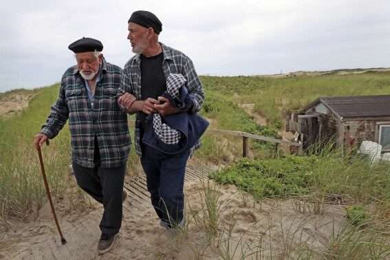 Artist Salvatore Del Deo walked around the shack, right rear, with the aid of his son Romolo Del Deo, on June 27, 2023, in Provincetown, Mass. The 95-year-old painter and his family who were threatened with eviction from their dune shack at the Cape Cod National Seashore in Mass., have won a reprieve. Lawyers for the painter worked out a deal with federal officials allowing Del Deo and his family to continue living in the shack for another five years. (David L Ryan/The Boston Globe via AP)