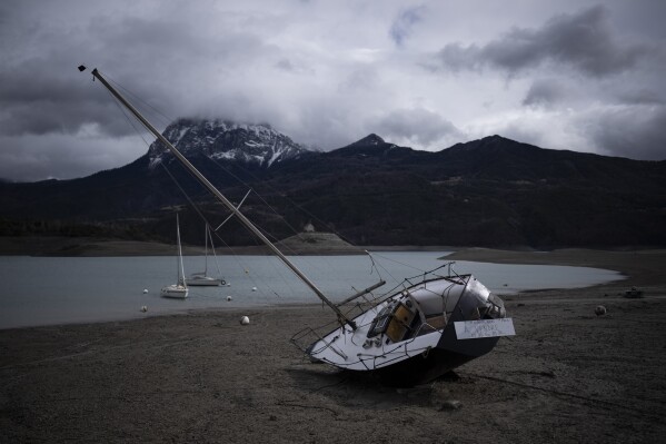 A stranded sail boat with a sign reading "for sale" lies above the water line at the Serre-Poncon resevoir in France, Tuesday, March 14, 2023. Human-caused climate change is lengthening droughts in southern France, meaning the reservoirs are increasingly drained to lower levels to maintain the power generation and water supply needed for nearby towns and cities. (AP Photo/Daniel Cole)
