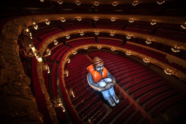 Sitting alone in a sea of empty red velvet chairs, a 6-meter-tall giant 'Inflatable Refugee' is photographed at Barcelona's Gran Teatre de Liceu opera house, Spain, Wednesday, June 12, 2024. Created by Belgian visual artists Schellekens & Peleman in 2015 following the refugee crisis in Europe by, they hope it to spark debate around the plight of refugees. The artistic intervention coincided with an alarming record: 120 million people around the world have been forcibly displaced from their homes due to conflict and other protracted crises, the U.N. Refugee announced on Thursday. (AP Photo/Emilio Morenatti)
