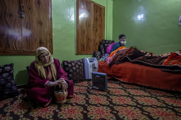 Abdul Rasheed, who suffers from chronic obstructive pulmonary disease (COPD), uses a portable oxygen concentrator while sitting on a bed as his wife Nora Rasheed warms herself with a traditional fire port called Kanger in downtown area of Srinagar, Indian controlled Kashmir, Saturday, Dec. 30, 2023. This year the unscheduled power outages, sometimes lasting 12 to 16 hours, have disrupted patient care for those with respiratory illnesses and affected businesses. (AP Photo/Dar Yasin)