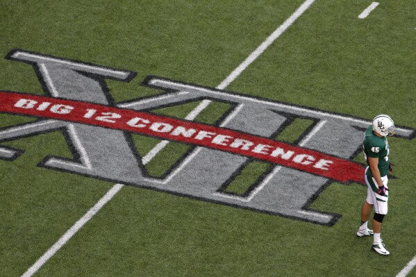 FILE - Baylor's Matt Ritchey stands by the Big 12 Conference logo on the field during the team's NCAA college football game against TCU in Waco, Texas, Oct. 13, 2012. The NCAA and the nation's five biggest conferences have agreed to pay nearly $2.8 billion to settle a host of antitrust claims,a monumental decision that sets the stage for a groundbreaking revenue-sharing model that could start directing millions of dollars directly to athletes as soon as the 2025 fall semester. (AP Photo/Tony Gutierrez, File)