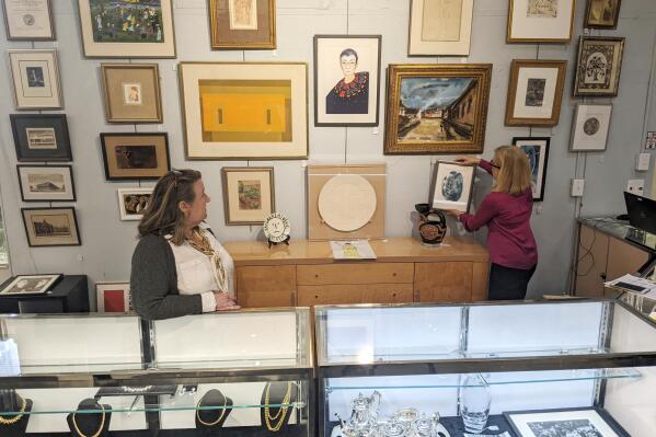 In this image from video, Elizabeth Haynie Wainstein, right, hangs up a piece of modern art belonging to the late Justice Ruth Bader Ginsburg inside Potomack Company Auctions in Alexandria, Va., Monday, April 11, 2022. (AP Photos/Nathan Ellgren)