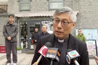 In this image from a video shot by Hong Kong broadcaster TVB, Hong Kong Bishop Stephen Chow talks to media during his visit to Beijing, with unidentified men standing behind him, on Thursday, April 20, 2023. Chow said in a historic trip to Beijing that he hopes the dioceses of the two cities will have more exchanges and cooperation, local media reported Thursday amid signs of Sino-Vatican strains. (TVB via AP)