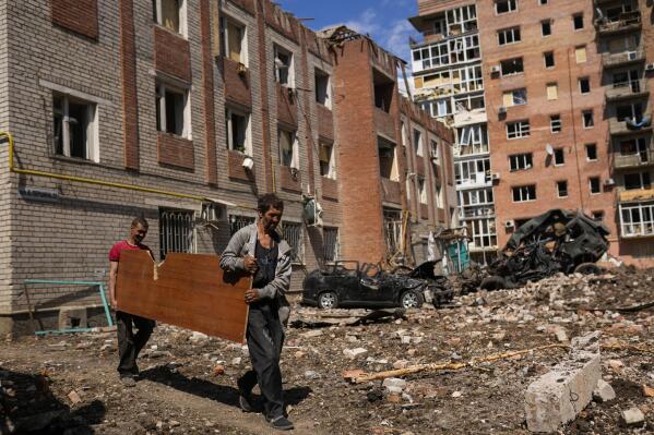 Two men carry a wooded panel next to heavily damaged buildings and destroyed cars in a Russian bombing in Bakhmut, eastern Ukraine, Tuesday, May 24, 2022. The town of Bakhmut has been coming under increasing artillery strikes, particularly over the last week, as Russian forces try to press forward to encircle the city of Sieverodonetsk to the northeast. (AP Photo/Francisco Seco)