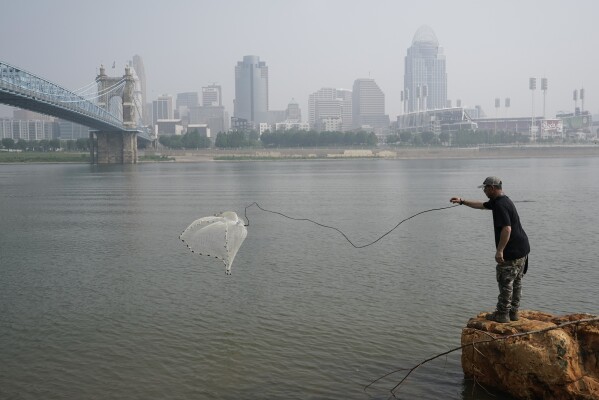 FILE - A man tosses a fishing net into the Ohio River as smoke from wildfires is visible over downtown Cincinnati, June 28, 2023. Forecasters say there won’t be large breaks for much of America anytime soon from eye-watering dangerous smoke from fire-struck Canada. (AP Photo/Joshua A. Bickel, File)