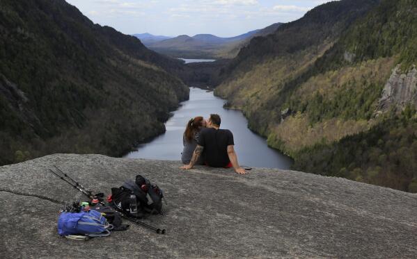 Sidney Gleason, left, and Joe Gorsuch, both of Syracuse, kiss while taking in the view of Lower Ausable Lake at Indian Head summit inside the Adirondack Mountain Reserve, Saturday, May 15, 2021, near St. Huberts, N.Y. A free reservation system went online recently to control the growing number of visitors packing the parking lot and tramping on the trails through the private land of the Adirondack Mountain Reserve. The increasingly common requirements, in effect from Maui to Maine, offer a trade-off to visitors, sacrificing spontaneity and ease of access for benefits like guaranteed parking spots and more elbow room in the woods. (AP Photo/Julie Jacobson)