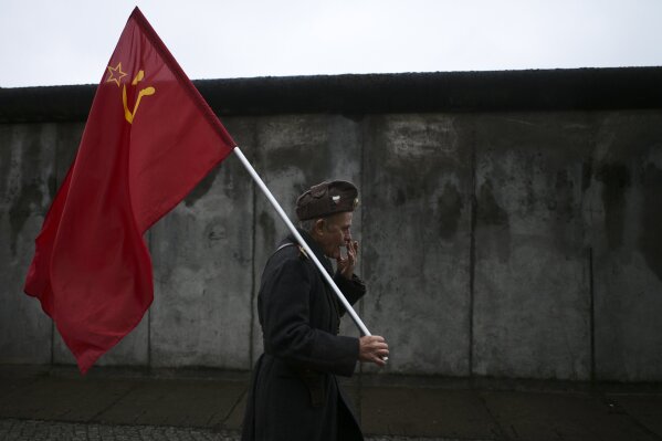 An elderly man with a Soviet flag walks in front of remains of the Berlin Wall after commemorations celebrating the 30th anniversary of the fall of the Berlin Wall at the Wall memorial site at Bernauer Strasse in Berlin, Saturday, Nov. 9, 2019. The man, who would not give his name, walks around with the flag as an protest against capitalism. (AP Photo/Markus Schreiber)