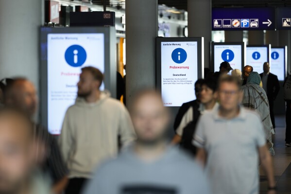 People walk in front of a screens displaying a nation wide test alarm message at the main train station in Berlin, Germany, Thursday, Sept. 14, 2023. Warning messages were blaring on cell phones, public displays and deafening alarms sounded across Germany in a nationwide test alert. The screen reads: "Official Warning- Test Alarm Germany There is no need for action Report your observations at https://warntag-umfrage.de." (AP Photo/Markus Schreiber)