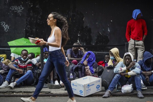Pedestrians pass migrants waiting in a queue outside of The Roosevelt Hotel that is being used by the city as temporary housing, Monday, July 31, 2023, in New York. (AP Photo/John Minchillo)