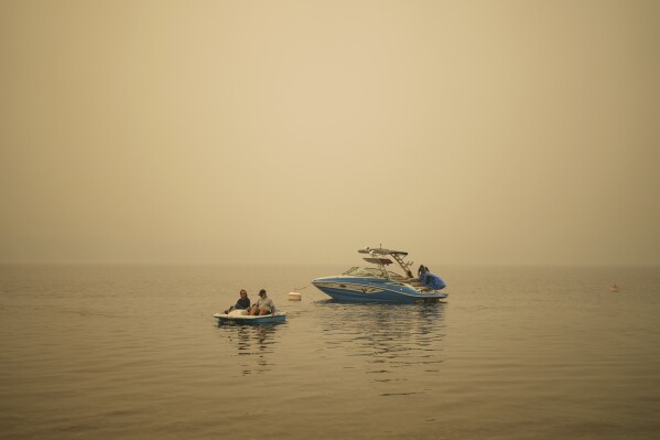 Smoke fills the air as Pat Manzuik and her husband Trevor use a paddleboat to get to shore after being given a boat ride by good samaritan Christy Dewalt, back right, back to their home they were evacuated from due to the Lower East Adams Lake wildfire, in Scotch Creek, Canada, Sunday, Aug. 20, 2023. (Darryl Dyck/The Canadian Press via AP)