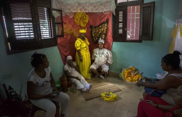 FILE - Marisa Ramirez Gutierrez, standing center, holds still on her symbolic throne dressed in the robe of the the "Iyawo," or bride, alongside a pregnant woman who represents birth, and her mother, who represents a woman who has already given birth, inside her home as part of many initiation ceremonies marking the start of her one-year journey to become a Yoruba priestess in Havana, Cuba, Sept. 11, 2015. Santería was born as a form of quiet resistance among Cuba’s Black communities. (AP Photo/Ramon Espinosa, File)