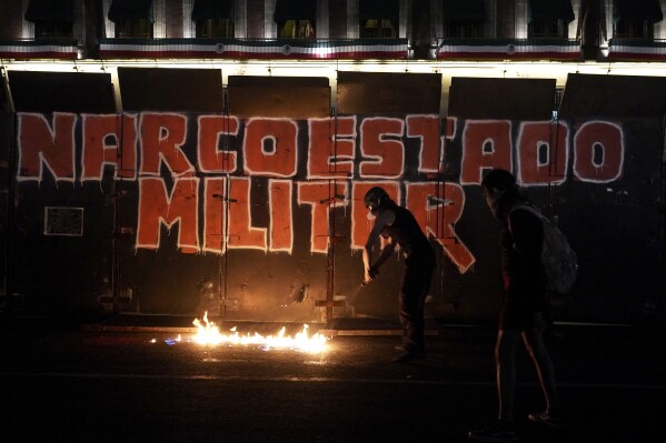 Demonstrators start a fire at the foot of the barrier protecting the National Palace during a march to commemorate the 9th anniversary of the disappearance of 43 missing Ayotzinapa university students, in Mexico City, Tuesday, Sept. 26, 2023. The graffiti on the barrier reads "Military Narcostate" in Spanish. (AP Photo/Marco Ugarte)