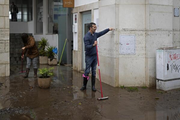 A man sweeping water from a building shows passers by the height that the water reached when the street was flooded overnight in Alges, just outside Lisbon, Tuesday, Dec. 13, 2022. An Atlantic storm slammed into the Iberian peninsula, leaving behind a trail of destruction Tuesday, especially in the Portuguese capital Lisbon, before moving eastward into Spain. (AP Photo/Armando Franca)