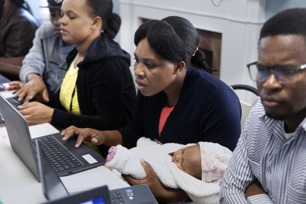 Jimene Admettre, center, sits next to her husband Ernseau, right, and holds her daughter Gabyana while learning computer skills, Friday, Dec. 22, 2023, in a rectory building where they are staying at the Bethel AME Church in the Jamaica Plain neighborhood of Boston. Demand for shelter has increased as Massachusetts struggles to find newly arriving migrants places to stay after hitting a state-imposed limit of 7,500 families in its emergency homeless shelter system last month. (AP Photo/Michael Dwyer)