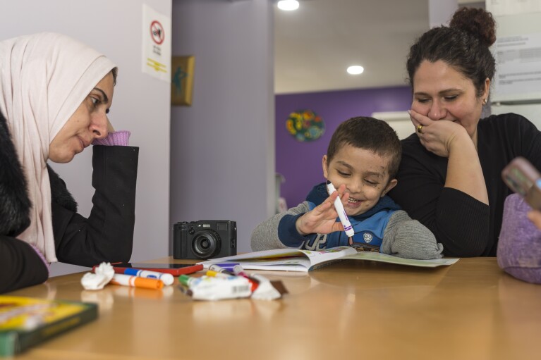 Four-year-old Omar Abu Kuwaik, center, works on activities in a math workbook with clinical social worker Sandy Lulu, right, as his aunt Maha Abu Kuwaik looks on, at the Global Medical Relief Fund residence, Sunday, Feb. 11, 2024, in the Staten Island borough of New York. Omar Abu Kuwaik is far from his home in Gaza. The 4-year-old's parents and sister were killed by an Israeli airstrike, when he lost part of his arm. (AP Photo/Peter K. Afriyie)