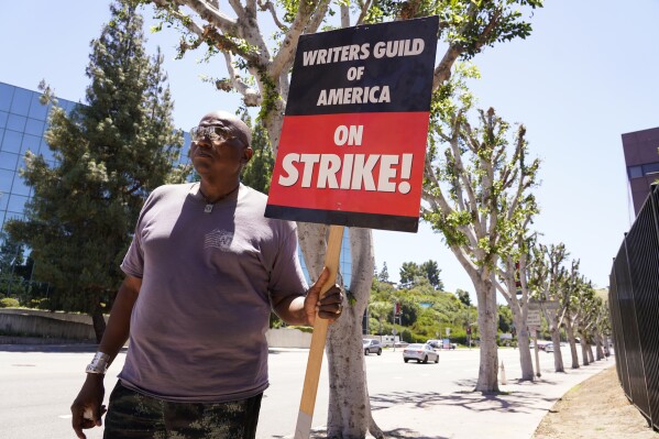 WGA and SAG-AFTRA member Dee Thompson walks past trees as he carries a sign on a picket line outside Universal studios on Wednesday, July 19, 2023, in Burbank, Calif. The actors strike comes more than two months after screenwriters began striking in their bid to get better pay and working conditions. (AP Photo/Chris Pizzello)