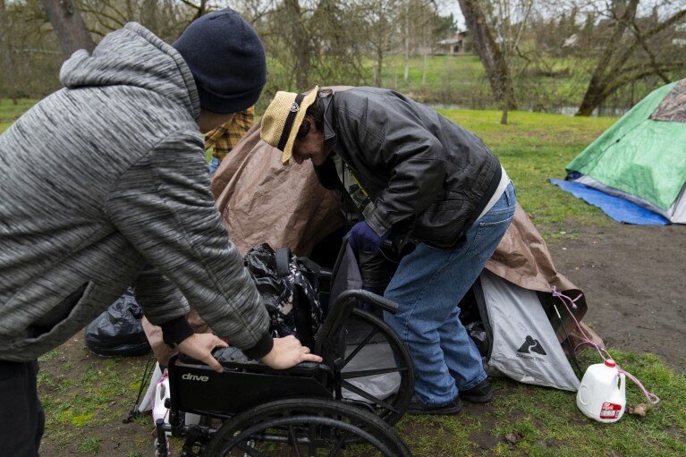 A volunteer holds on to a wheelchair as they help Max Hartfelt into his tent after relocating him from one park to another on Saturday, March 23, 2024, in Grants Pass, Ore. (AP Photo/Jenny Kane)