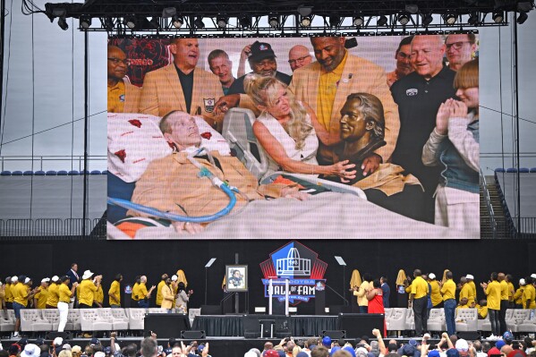 Misty McMichael, wife of former NFL player Steve McMichael, left, unveils his bust during an induction ceremony from their home, on the video screen at the Pro Football Hall of Fame, in Canton, Ohio, Saturday, Aug. 3, 2024. (AP Photo/David Richard)