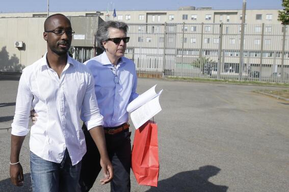 FILE - Rudy Guede, left, is greeted by an unidentified person as he leaves the penitentiary for a temporary release of thirty-six hours, in Viterbo, Italy, on June 25, 2016. Guede, the only person convicted in the 2007 murder of British student Meredith Kercher has been freed after serving most of his 16-year prison sentence. Italian news agencies LaPresse and ANSA quoted attorney Fabrizio Ballarini as saying Rudy Guede’s planned Jan. 4 release had been moved up a few weeks by a judge and was freed on Tuesday.(AP Photo/Gregorio Borgia)