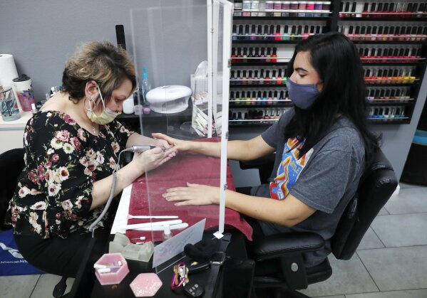 FILE - In this April 24, 2020, file photo, manicurist Rhonda Simpson, left, polishes nails for her customer Faith at the reopened Salon A la Mode in Dallas. The salon installed a barrier between the two to avoid the spread of COVID-19. As some governors across the United States begin to ease restrictions imposed to stop the spread of the coronavirus, hopes are soaring that life as we knew it might be returning. But the plans emerging in many states indicate that “normal” is still a long way off. (AP Photo/LM Otero)