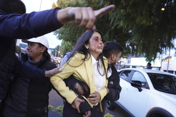 A bloodied woman is carried away after presidential candidate Fernando Villavicencio was shot to death at a campaign rally outside a school in Quito, Ecuador, Wednesday, Aug. 9, 2023. Villavicencio, 59, who was known for speaking up against drug cartels, was assassinated less than two weeks before a special presidential election. (API via AP)