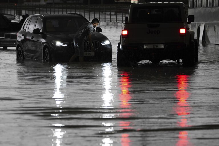 A man tries to work on his stalled SUV in standing water in Dubai, United Arab Emirates, Tuesday, April 16, 2024. Heavy rains lashed the United Arab Emirates on Tuesday, flooding out portions of major highways and leaving vehicles abandoned on roadways across Dubai. Meanwhile, the death toll in separate heavy flooding in neighboring Oman rose to 18 with others still missing as the sultanate prepared for the storm. (AP Photo/Jon Gambrell)