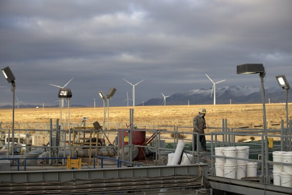 A rig operator walks through a Fervo Energy geothermal drilling site near Milford, Utah Sunday, Nov. 26, 2023, with a wind farm in the background. In Nevada, Fervo Energy's first operational geothermal project has begun pumping carbon-free electricity onto that state's electric grid to power Google data centers, Google announced Tuesday, Nov. 28. That pilot will help Fervo launch projects like this one in Utah to deliver far more carbon-free electricity to the grid. (AP Photo/Ellen Schmidt)