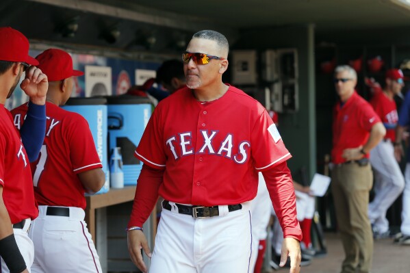 FILE -Texas Rangers first base coach Hector Ortiz (4) walks through the dugout talking with players during a baseball game against the Los Angeles Angels on Sunday, Sept. 3, 2017, in Arlington, Texas. Hector Ortiz, who spent the past 18 years as a manager and coach in the Texas Rangers organization, died Wednesday, Feb. 28, 2024 after a long battle with cancer. He was 54.(AP Photo/Tony Gutierrez, File)