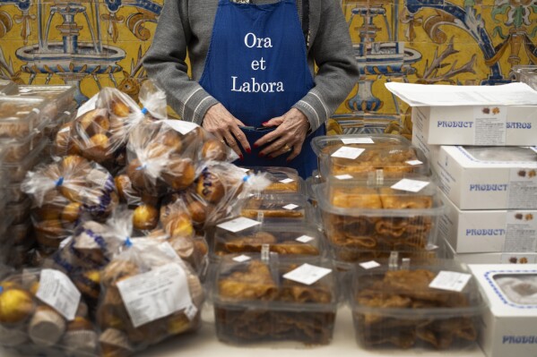 A volunteer, wearing an apron which reads, "Pray and Work" in Latin, waits for customers to sell cakes made by cloistered nuns, at a market at the Reales Alcazares in Seville, Spain, on Tuesday, Dec. 5, 2023. It's the fortnight before Christmas and all through the world's Catholic convents, nuns and monks are extra busy preparing the traditional delicacies they sell to a loyal fan base even in rapidly secularizing countries. (AP Photo/Laura Leon)