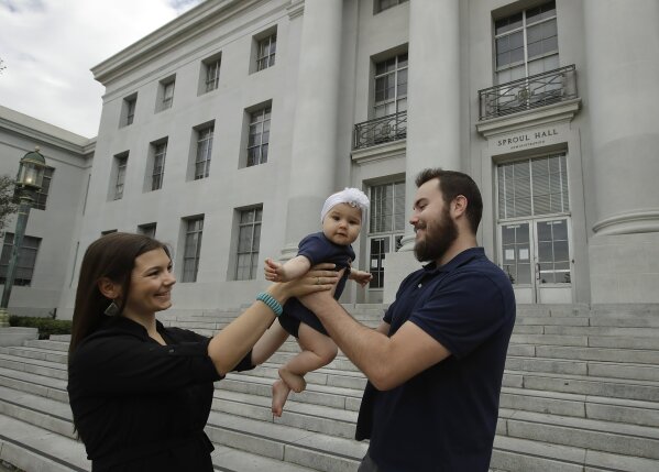University of California at Berkeley graduate Tyler Lyson, right, reaches for his 8-month-old daughter, Lisa, from his wife, Lucie, in front of Sproul Hall on the closed Cal campus in Berkeley, Calif., on Monday, May 11, 2020. As he struggles to find work, Tyler Lyson is considering leaving Berkeley to move back home to Post Falls, Idaho, where it’s cheaper, even though it would feel like giving up on his dreams. (AP Photo/Ben Margot)