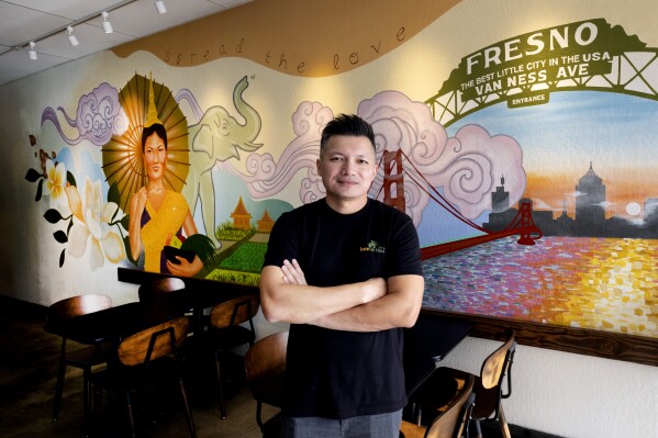 David Rasavong stands by a mural depicting his family's journey from Laos to San Francisco and then to Fresno, in his restaurant "Love & Thai" in Fresno, Calif. on Wednesday, Dec. 20, 2023. Rasavong's body still tenses up when recounting how a so-called animal welfare crusader in May implied on social media that his 7-month-old restaurant, Tasty Thai, owned a pitbull tied up at a home next door. What's more, the dog would eventually be meal fodder. By the next day, vitriolic comments, voicemails and calls rained down. (AP Photo/Richard Vogel)