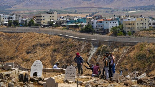 Residents dig a grave at a cemetery on the Lebanese side of the Lebanese-Israeli border in the southern village of Wazzani with the walled northern part of Ghajar village in the background, Lebanon, Tuesday, July 11, 2023. The little village of Ghajar has been a point of contention between Israel and Lebanon for years, split in two by the border between Lebanon and the Israeli-occupied Golan Heights. The dispute has begun to heat up again.(AP Photo/Hassan Ammar)