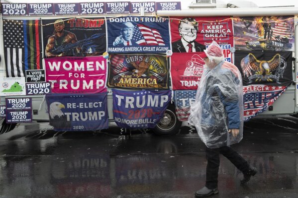 A man in a rain cover walks past a vehicle decorated with flags in support of President Donald Trump near the venue where  the president will hold a campaign rally in the evening, Monday, Feb. 10, 2020, in Manchester, N.H. (AP Photo/Mary Altaffer)