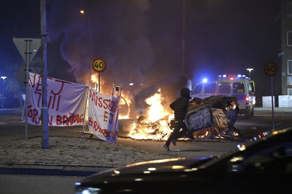 A person runs past a burning car and a banner reading, "No to Quran burning. Don't abuse freedom of expression" placed on the roundabout, at Ramel väg, in Malmo, Sweden, early Monday Sept. 4, 2023. Clashes in an immigrant neighborhood in Sweden’s third largest city after an anti-Muslim protester set fire to the Quran with police being pelted with rocks and dozens of cars set on fire. Police said Monday, adding the tensions now have died down. (Johan Nilsson /TT News Agency via AP)